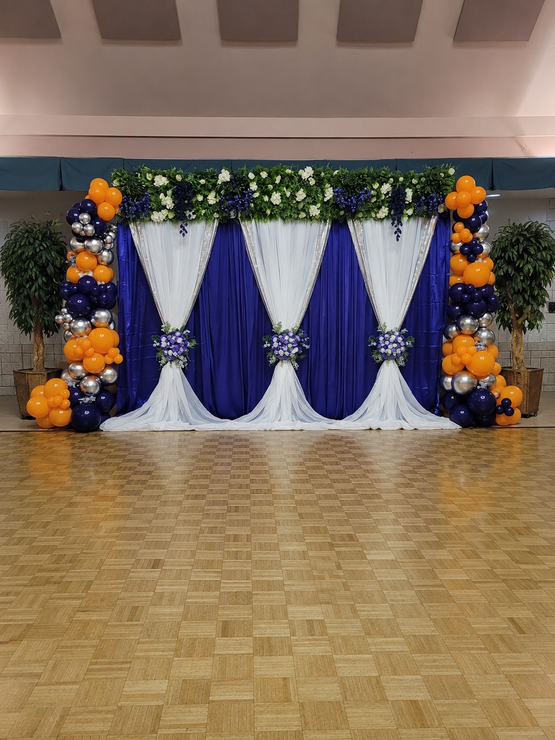 Royal blue and white draped stage backdrop with florals and orange, navy and silver balloon columns